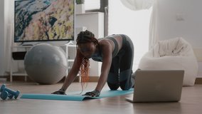 Trainer woman with black skin practicing fitness position during yoga morning workout sitting on aerobic map in living room. Adult working body flexibility watching gymnastics online video on laptop