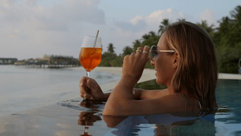 Young woman having a drink in an infinity pool at sunset. Female enjoying a tropical vacation in a luxury resort in the Maldives 