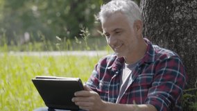 Man relaxing by a tree in country field, using digital tablet