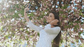 Young smiling woman in summer white dress taking selfie self portrait photos on smartphone. Model posing on park sakura trees background. Female showing positive face emotions.