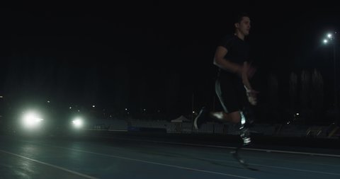 Cinematic shot of young man with disability with legs prosthesis running with effort with racing auto behind on car track at night. Concept of persons with disabilities active lifestyle, determination
