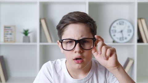 Afraid and shocked boy 13 years old in white shirt takes off glasses and cover mouth with his hand. Surprised kid teenager scared of bad news looking to the camera on home interior background.