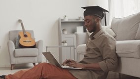Lockdown of young African-American man wearing graduation hat sitting on floor at home with laptop on knees, finishing online graduation ceremony, taking hat off and typing on keyboard