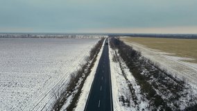 Drone shoots video of car and truck drive on rural paved road with snow on both sides in winter between agricultural fields. Snowy frozen highway