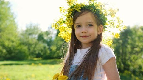 Portrait of a cute little girl in a flower wreath on her head with a dandelion in her hands, she looks at the camera against the background of a sunset with sun glare