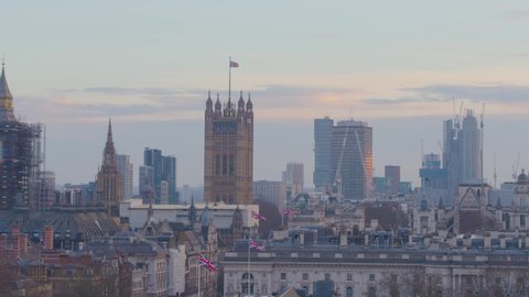 Aerial drone view tracking fly past shot looking towards Big Ben and Parliament skyline at sunset in London, with Union Jack flags flying in wind