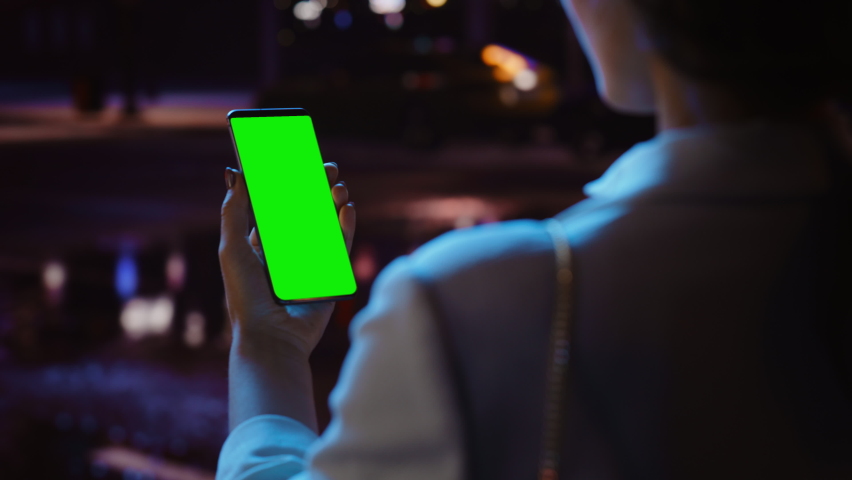 Woman using Chroma Key Smartphone while Standing, Looking at the Green Screen. Night City Street Full of Neon Light. Female Using Mobile Phone. Over the Shoulder Closeup Tracking Shot Focus on Display