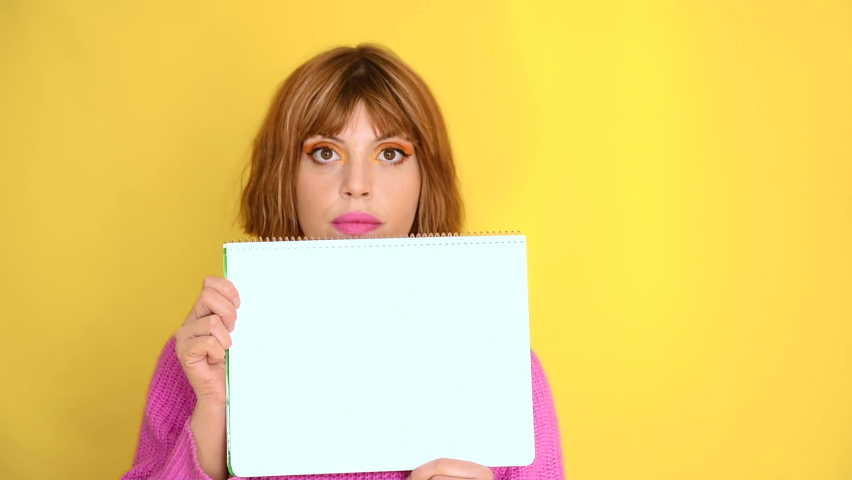 A girl standing in front of a yellow background with short hair and a pink sweater, she is holding a white notebook, she is looking intently at the notebook, she is looking for approval.