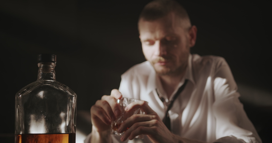 Alcohol deprives us of willpower, it gives a false sense of relaxation and freedom. A man is struggling with alcohol addiction. He's sitting in front of a bottle of whiskey and holding a glass of whiskey. | Shutterstock HD Video #1072963019