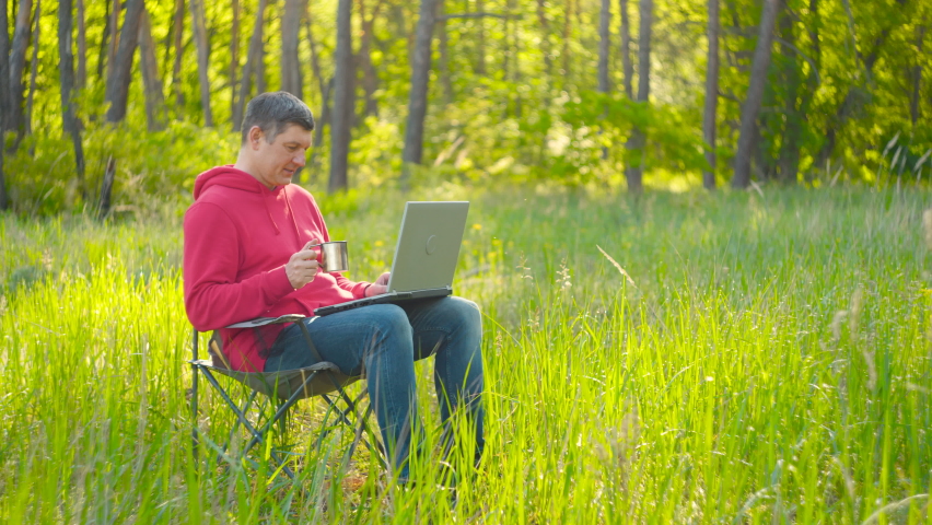 Man working on laptop in summer forest At The Sunny Day. Busy freelance worker drinking tea and working on modern technology laptop. Freelancer Remote Work Concept. 4K