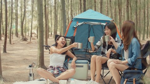Group of young asia camper friends sitting in chairs by tent in forest. Teenager girl traveler relax and talk on a summer day at campsite. Outdoor activity, adventure travel, or holiday vacation.