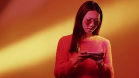 Mobile gaming. Color light girl. Online tournament. Cyber technology. Pink neon excited female Asian gamer playing on phone isolated on glowing yellow orange gradient background.