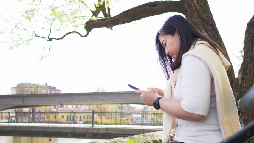 Asian black straight hair woman sitting by the river and using her phone to have a chat with friend, Looking up and seeing many birds on the sky. Beautiful day