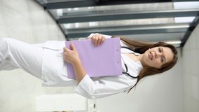 Happy young caucasian woman doctor wearing white medical coat and stethoscope with documents in her hands looking at camera. Smiling female doctor or nurse in hospital office. Vertical video