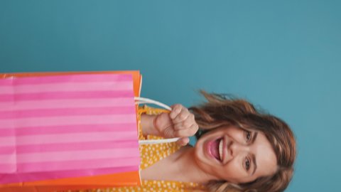 Vertical video Happy Young Woman Shopping Bags Dancing in front Rhythmically Moves her Arms Up Down, Looks Camera on Blue Background Fashion. Sale in Malls. Black Friday. Lifestyle
