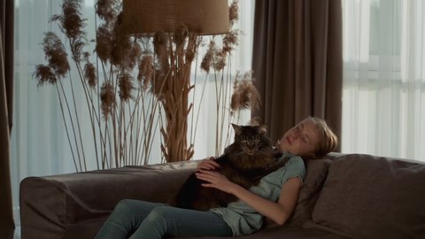 A cute teenage girl petting her pet while sitting on the sofa in the living room. Beautiful cat, Maine Coon breed. Medium shot, Slow motion.