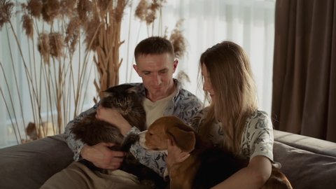 A happy young couple of European appearance hug their pets sitting on the sofa in the living room. A beautiful dog of the Beagle breed and pretty Maine coon cat. Slow motion