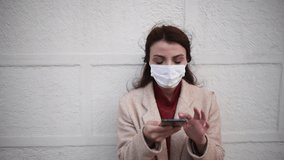 Slow Motion: Beautiful girl wearing protective medical mask and fashionable clothes speaks with a smart phone in front of wall. New normal lifestyle concept.