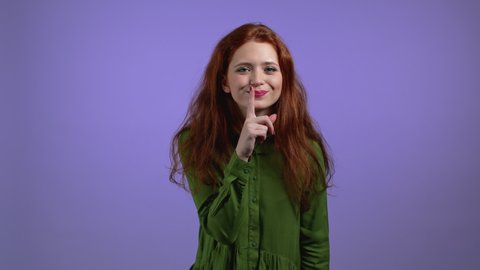 Smiling woman holding finger on her lips over violet background. Gesture of shhh, secret, silence. Close up