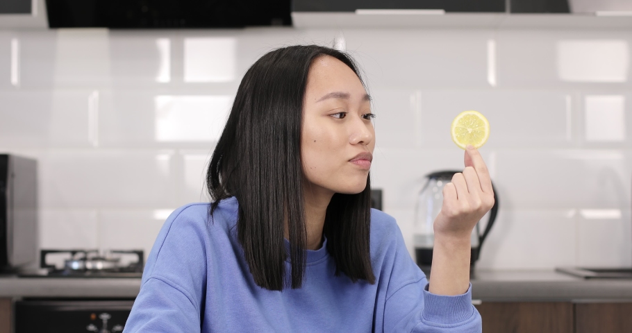 Attractive Asian woman showing lemon wedges in the kitchen. A young girl sits at a table and eats a sour fruit.