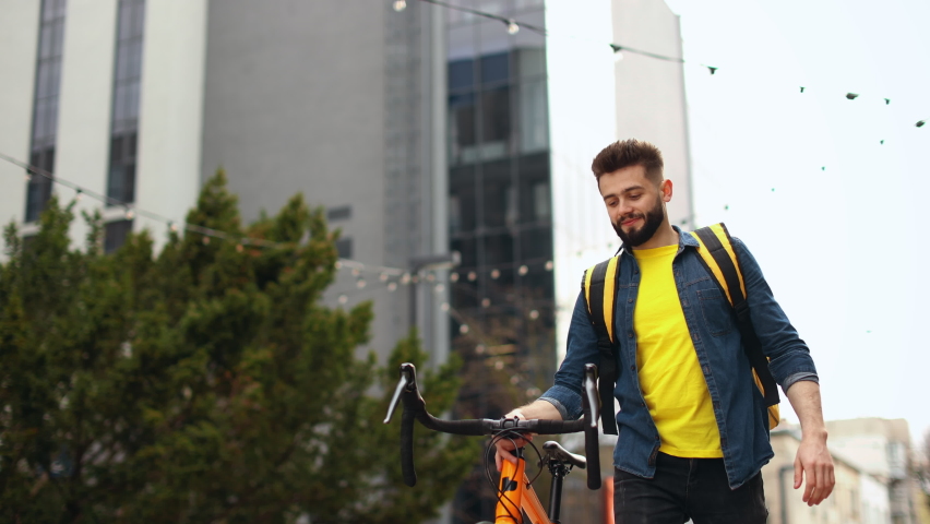 The food delivery guy is going and smiling. He is looking ahead. He is holding a bicycle and carrying a large backpack on his shoulders. The camera is shooting from below. Portrait shooting. 4K