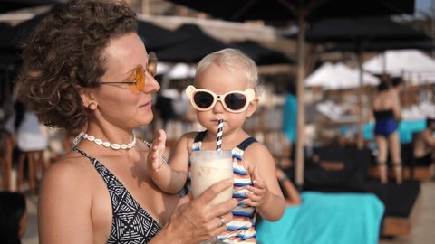 Young Caucasian mother holds her baby girl drinking a milkshake from glass with a paper straw. Kids friendly beach party