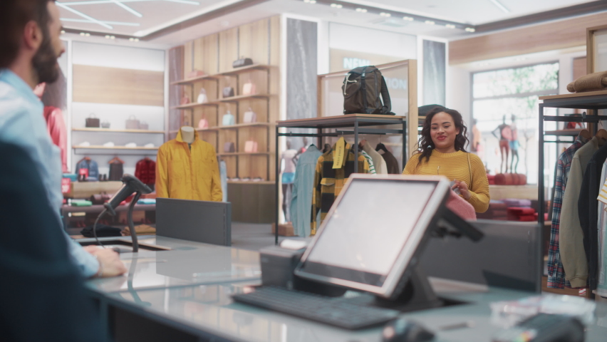 Clothing Store Checkout Cashier Counter: Woman and Male Retail Sales Managers Accept NFC Smartphone and Credit Card Payments from a Young Female Customers for Clothes and Pack Them in Recyclable Bags. Royalty-Free Stock Footage #1073014880