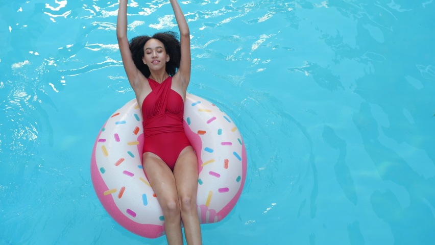 Ethnicity curly female floating in blue water in hotel pool sitting in inflatable tube raises hands above her and smile looking at camera. Afro student girl relax in tourist complex swimming on circle Royalty-Free Stock Footage #1073015120