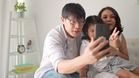 Happy Asian family spending time together at home. Parents using smartphone taking selfie or video call in living room. Father and mother with child girl kid enjoy and having fun weekend activity