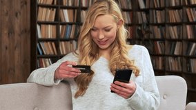 Young blonde woman sitting on the couch and makes purchases online using credit card and smart phone close-up