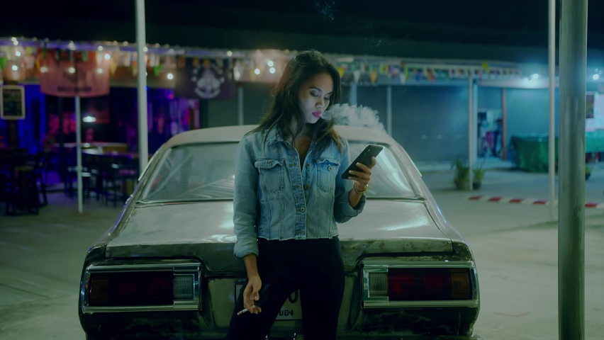 A girl smokes in front of a rusty classic car while typing a message late at night