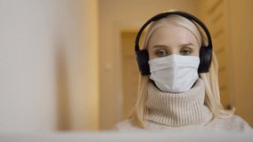 A young blonde woman in headphones and a medical mask communicates by video call via a laptop. Remote work during a pandemic. 4K UHD