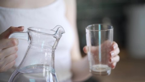 Beautiful young woman pours water into a glass and drinks close-up. The blonde pours water from a transparent decanter.
