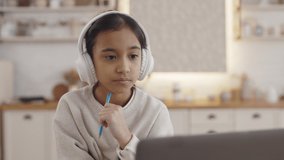Portrait of pretty pupil in wireless headphones staying at home and studying online. Focused indian girl with dark hair using laptop for education.