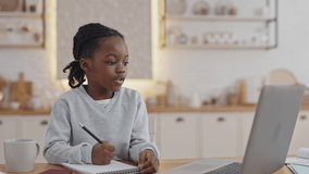 Cute little pupil using wireless laptop for video lesson while sitting at kitchen table. African american child greeting teacher by waving hand.