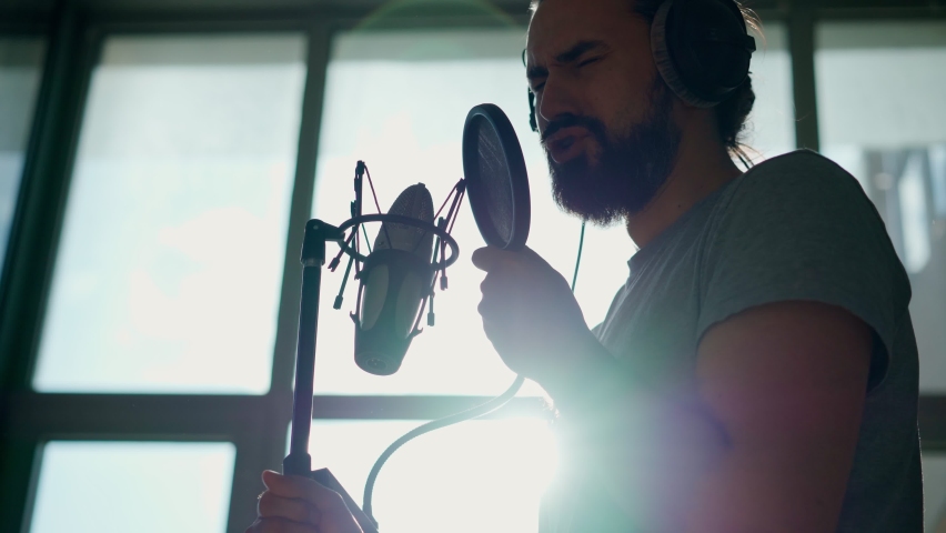 A young bearded man with headphones sings into a microphone on a stand with a pop filter, singer records a song at a recording studio.