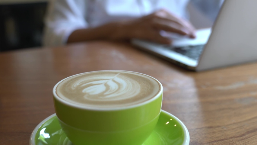 Person working on his laptop at the background with selective focus on cup of coffee on table. Working in a coworking office or café. Distant or remote work concept.