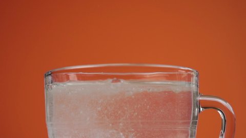 Effervescent soluble pill falls into a glass cup of clean drinking water with a splash and dissolves with bubbles. Slow motion. Close up. On a colored orange background
