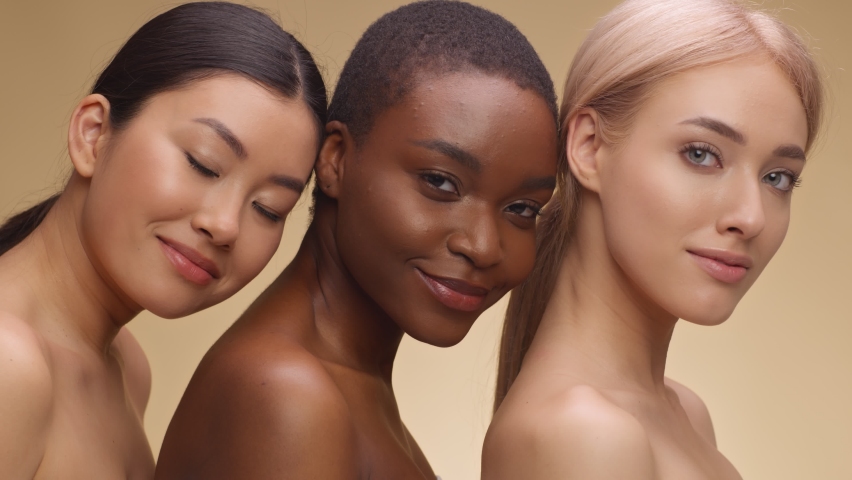 Multiethnic beauty. Fashion portrait of three diverse ladies with bare shoulders posing together and smiling to camera over beige studio backgroound Royalty-Free Stock Footage #1073065352