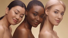 Multiethnic beauty. Fashion portrait of three diverse ladies with bare shoulders posing together and smiling to camera over beige studio backgroound