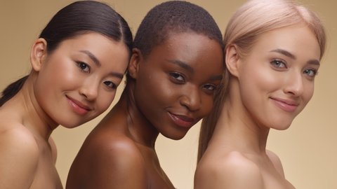 Multiethnic beauty. Fashion portrait of three diverse ladies with bare shoulders posing together and smiling to camera over beige studio backgroound