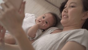 asian woman mother parent using mobile smart phone taking selfie photo with baby infant on bed at home. family video call via smartphone.  