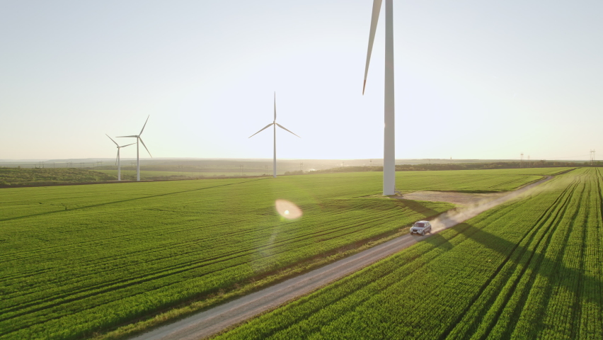 Car drives along rural road kicking up dust among green wheat field aerial view. Large wind turbines against blue sky at sunset in spring. Wind park agricultural farm. Alternative energy eco nature Royalty-Free Stock Footage #1073075045