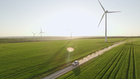 Car drives along rural road kicking up dust among green wheat field aerial view. Large wind turbines against blue sky at sunset in spring. Wind park agricultural farm. Alternative energy eco nature