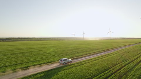 Car drives along rural road kicking up dust among green wheat field Large wind turbines against blue sky at sunset in spring. Wind park and agricultural farm. Alternative energy eco nature concept