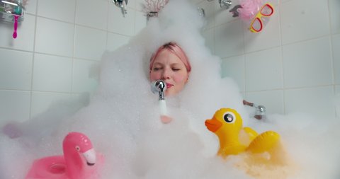 Funny woman sits in bath filled with foam, holds shower singing songs. Female in foam from head to toe in the bath fooling around