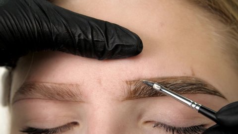 Makeup artist's hand in black gloves applies brow paste to the eyebrow. Attractive female face. Portrait of a beautiful young woman. Professional stylish permanent makeup, eyebrow tinting. Close up