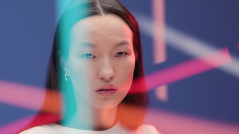 Abstract portrait of young asian woman in vibrant colorful light of bright neon lines. Female person looks at camera at blue background close-up. Face of adult girl with nude make-up in fashion studio