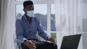 Young man in coronavirus face mask sitting in sunny home office conferencing online on laptop. Portrait of concentrated African American confident employee using video chat on Covid-19 lockdown