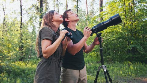 Birdwatchers. Young couple watching birds in the park. Man and woman use binoculars and camera with telephoto lens to spot birds in the summer park